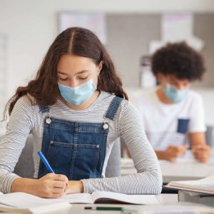 High school student taking notes from book while wearing face mask