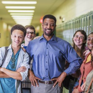 A multi-ethnic group of six high school students, 15 to 18 years old, hanging out together in the hallway by the lockers, between classes, with the teacher or school principal, a mature African-American man in his 50s. They are smiling at the camera.