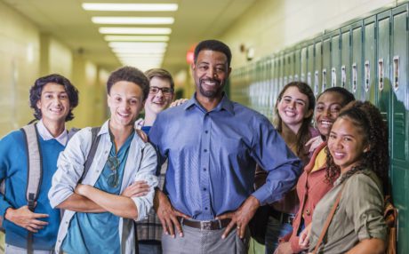 A multi-ethnic group of six high school students, 15 to 18 years old, hanging out together in the hallway by the lockers, between classes, with the teacher or school principal, a mature African-American man in his 50s. They are smiling at the camera.