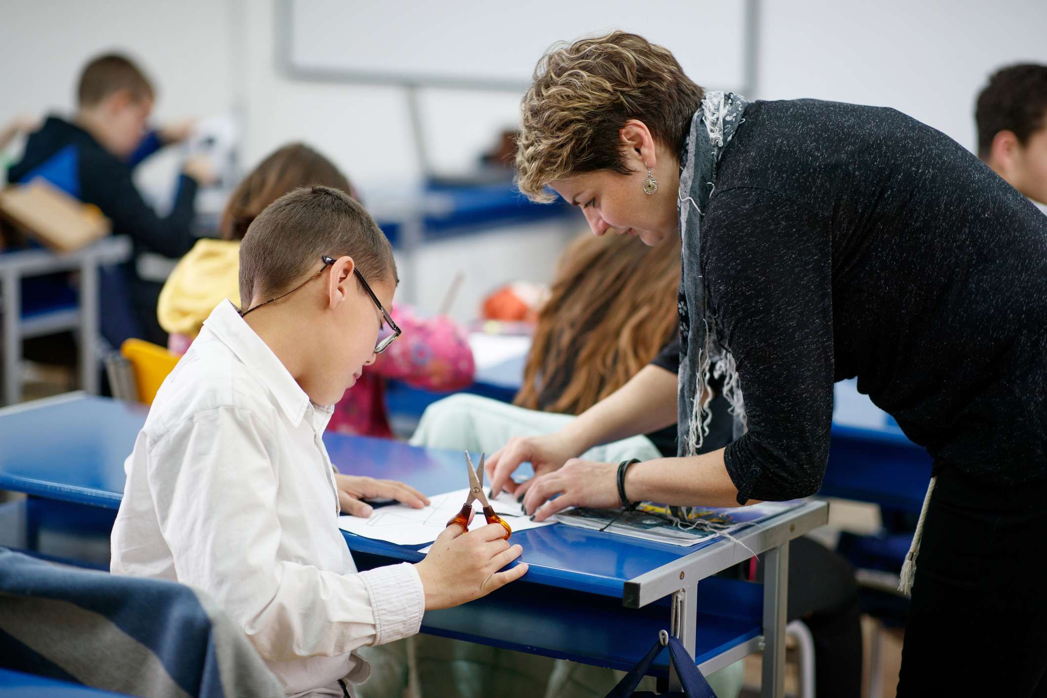 Female teacher working with a special need elementary age boy, she is directing him how to cut paper following pattern